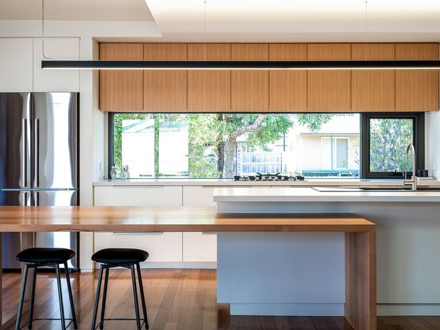 A bright contemporary kitchen with a long timber island and breakfast bar, two black stools, white lower cabinetry, warm timber overhead cabinetry, stainless-steel refrigerator, pale benchtops, and a large horizontal window overlooking greenery. A slim linear pendant light runs above the island, and timber flooring adds warmth.