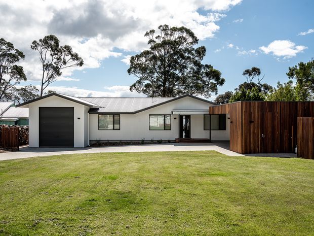 Single-storey contemporary residential house viewed from the front across a broad lawn. The home has a low-pitched metal gable roof, light-coloured horizontal cladding, dark-framed windows, a black garage door, and a recessed entry. A tall dark timber privacy fence forms a prominent side element, with mature trees behind the property under a bright partly cloudy sky.
