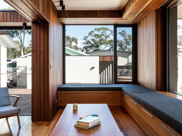 A warm, timber-lined built-in window seat and workspace nook in a contemporary home. Vertical timber slats cover the walls and ceiling, with a large black-framed window overlooking a courtyard and adjacent building. The built-in joinery includes deep drawers, a dark upholstered bench seat, and a small timber table with books, while a lounge chair sits beside glazed doors.