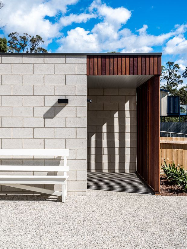 Exterior view of a contemporary residential entry or covered passage. The facade combines pale exposed concrete block masonry with a dark reddish-brown vertical timber-clad portal and soffit. A recessed walkway is shaded by the projecting timber-framed canopy, with a white built-in bench along the wall and a textured light aggregate paving surface. Strong sunlight creates crisp shadows, while a timber fence and planted garden are visible to the side.