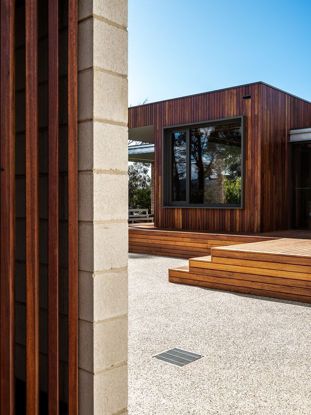 A close exterior view of a modern timber-clad building arranged around a paved courtyard. The facade features vertically oriented dark timber boards, large dark-framed windows, a simple pitched roofline, raised timber decking and broad timber steps. A pale textured masonry wall and vertical timber screening frame the foreground.