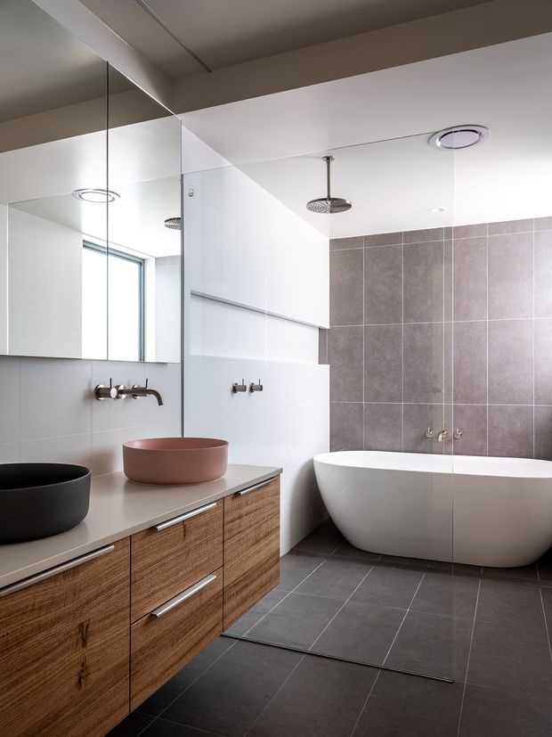 Contemporary bathroom with a freestanding white bathtub, large-format grey wall and floor tiles, frameless glass shower enclosure, wall-mounted chrome fixtures, and a timber vanity with a pale stone benchtop and vessel basins. A large mirror and minimal recessed lighting contribute to the clean, refined design.