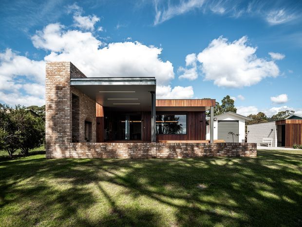 Exterior view of a contemporary single-storey residence set in a landscaped suburban or semi-rural site. The house combines exposed light brick walls, dark timber cladding, extensive glazing, and a projecting flat-roofed covered outdoor area. Separate garage and ancillary structures are visible to the right, with a broad lawn in the foreground under a bright blue sky.