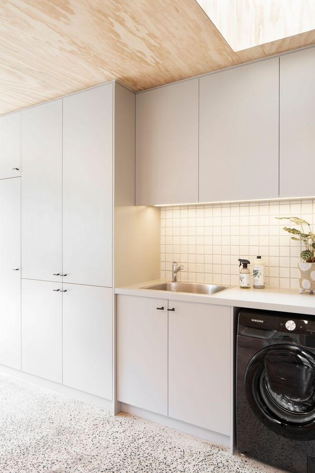 A compact, contemporary laundry with full-height pale cabinetry, integrated storage, a stainless-steel sink, washing machine, warm under-cabinet lighting, and a small square tiled splashback. The room features a light timber-lined ceiling and a terrazzo-style floor.