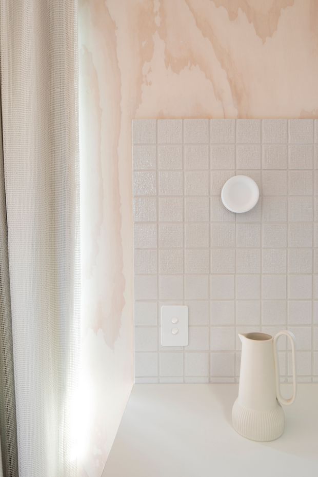 Close-up of a softly lit interior corner featuring a pale square tiled splashback, a white electrical outlet, and a white countertop with a sculptural ceramic pitcher. A sheer textured curtain hangs beside a softly mottled pale pink wall. The restrained palette and tactile finishes create a calm, minimalist kitchen or kitchenette setting.