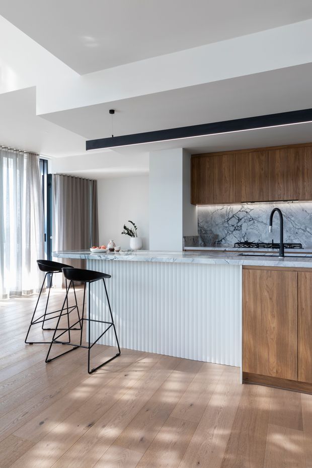 Bright contemporary kitchen with a long island and two black bar stools. The kitchen features warm timber cabinetry, a veined stone benchtop and splashback, a fluted white island front, black tapware and a slim linear pendant light. Full-height sheer curtains and pale timber flooring add an airy, refined feel.