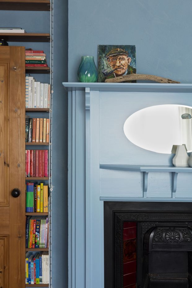 Close-up interior detail showing a traditional painted fireplace surround in pale blue, with a black ornate cast-iron fireplace insert and red tiled detailing. A built-in timber bookshelf filled with books sits beside it, while the mantel displays a green glass vase, artwork, and a natural wood object. The scene has a richly coloured, curated, heritage character.