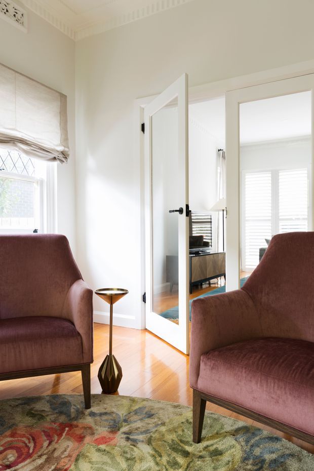 Bright, refined sitting room with two upholstered mid-century-style armchairs, a slim sculptural side table, patterned area rug, polished timber flooring, white walls, and ornate ceiling cornice detailing. Open mirrored French doors lead into an adjacent living area with a timber console, television, and plantation shutters. Natural light enters through the window and shutters.