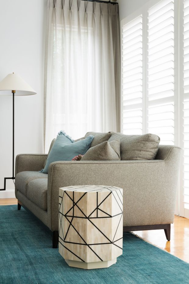 Bright, refined living room vignette featuring a large taupe upholstered sofa with pale blue and neutral cushions, a slender black floor lamp, full-height sheer curtains, and white plantation shutters. A geometric light-wood and black side table sits on a large teal rug over timber flooring, with abundant natural light.