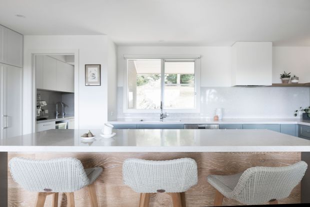 Bright contemporary white kitchen viewed from the dining side, featuring a large pale stone island with three woven bar stools, white cabinetry, a glossy light splashback, stainless steel sink and tap, and a window overlooking greenery. The space has a clean, airy, minimal appearance with warm timber detailing at the island base.