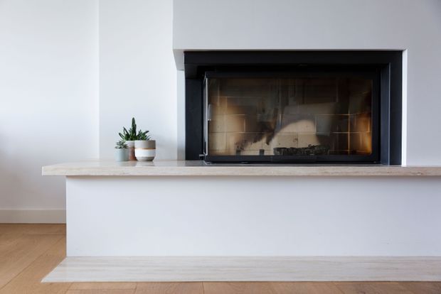Minimal contemporary fireplace feature wall with a black-framed built-in firebox set into a white plastered surround. A pale stone or concrete mantel projects across the wall above a simple white hearth, with a light timber floor and small potted plants adding subtle warmth.
