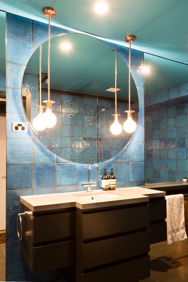A dramatic contemporary bathroom featuring glossy blue square wall tiles with contrasting grout, a large round mirror, twin white globe pendant lights with brass-toned stems, and a black floating vanity with a pale benchtop and integrated basin. A secondary bathtub is visible at the right.