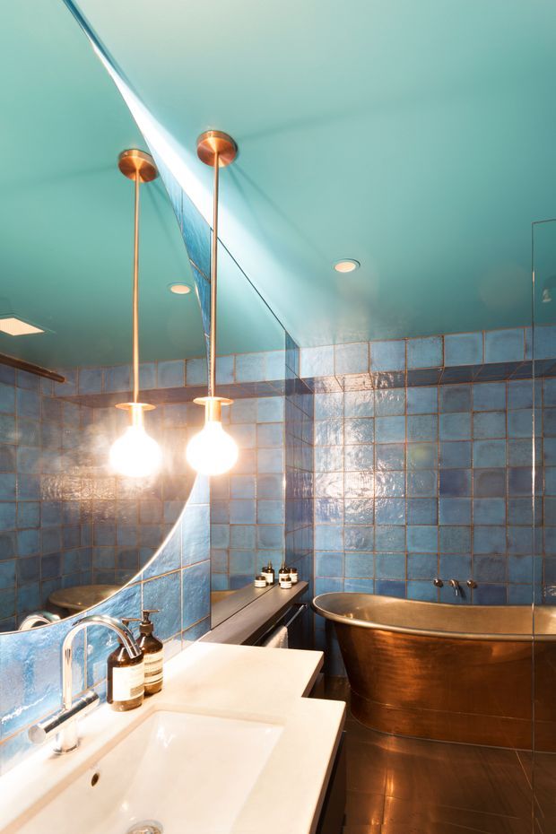 A compact, richly styled bathroom featuring glossy blue square wall tiles with dark grout, a teal painted ceiling, a large curved mirror, twin copper pendant lights, a white double vanity, and a freestanding metallic copper bathtub. The space has a dramatic, jewel-toned atmosphere with warm reflective lighting.