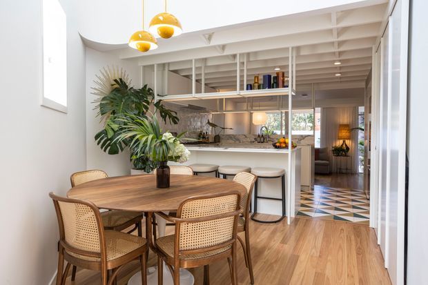 Bright open-plan dining and kitchen interior with a warm timber dining table and cane-back chairs in the foreground. The kitchen beyond features a marble-look island with stools, open shelving, glass-front storage, and abundant indoor plants. White exposed ceiling beams, timber flooring, curved walls, pendant lights, and patterned floor tiles in the hallway create a refined contemporary tropical feel.