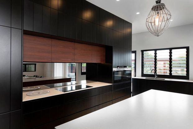 Contemporary kitchen with extensive dark cabinetry, warm timber overhead cabinetry, a pale stone island benchtop, integrated appliances, a mirrored splashback, and a black-framed louvred window above the sink. A sculptural wire pendant light provides a focal point over the island.