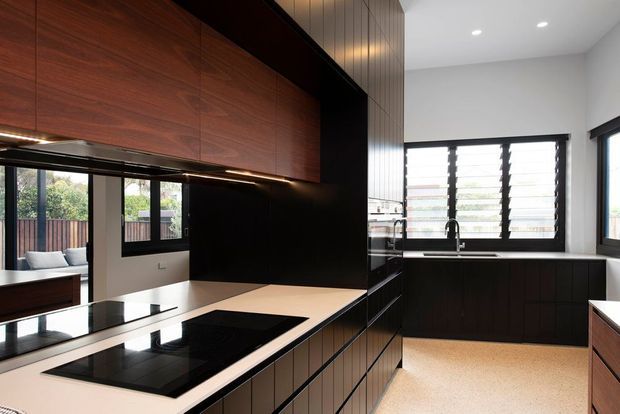 A contemporary galley-style kitchen with dark timber cabinetry, black integrated appliances and benchtops, a light stone or solid-surface island, and a large bank of louvred windows above the sink. Warm timber ceiling and cabinetry contrast with white walls and a pale speckled floor. A small outdoor area is visible through the glazing.