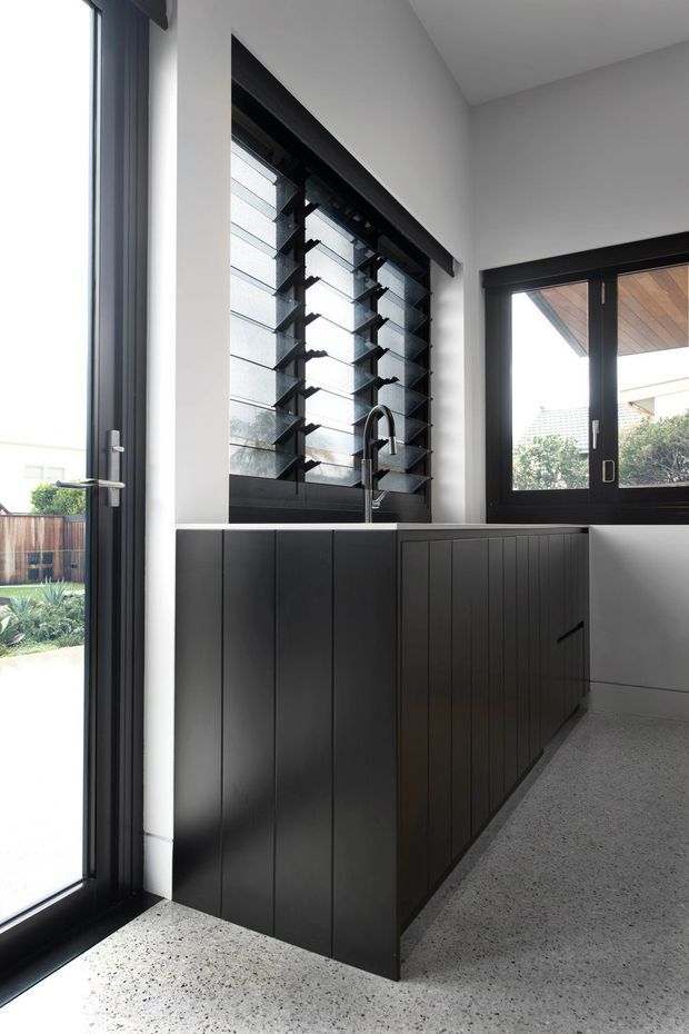 A bright contemporary kitchen with a compact black island featuring vertically grooved cabinetry and a dark integrated benchtop. A black-framed louvred window sits above the sink, with additional black-framed glazing and a glazed door bringing in abundant daylight. The floor is finished in light speckled terrazzo, while the walls and ceiling are white.