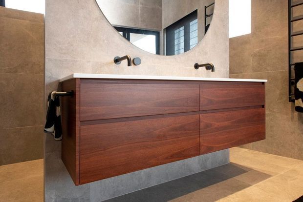 Contemporary bathroom featuring a floating dark timber double vanity with integrated drawers, a pale stone or tiled benchtop, wall-mounted black tapware, large-format warm neutral wall and floor tiles, and a broad rounded mirror. A window and reflected glazing provide natural light.