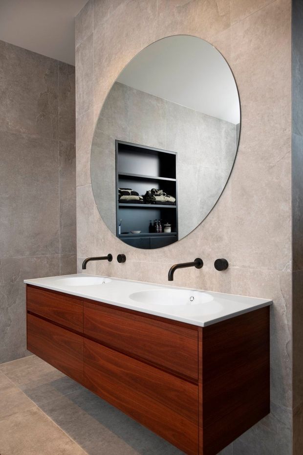 Contemporary bathroom featuring a large double-sink timber vanity with a white integrated benchtop, wall-mounted black tapware, an oversized arched mirror, and warm grey stone-look wall and floor tiles. A recessed open shelf is reflected in the mirror.