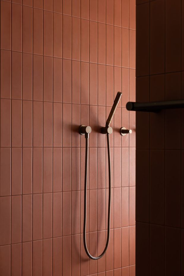 Close-up of a contemporary shower installation against a wall clad in narrow terracotta-pink rectangular tiles. The wall-mounted shower controls, handheld shower head, flexible hose, and horizontal dark metal fixture create a minimalist, warm-toned bathroom composition with dramatic side lighting.