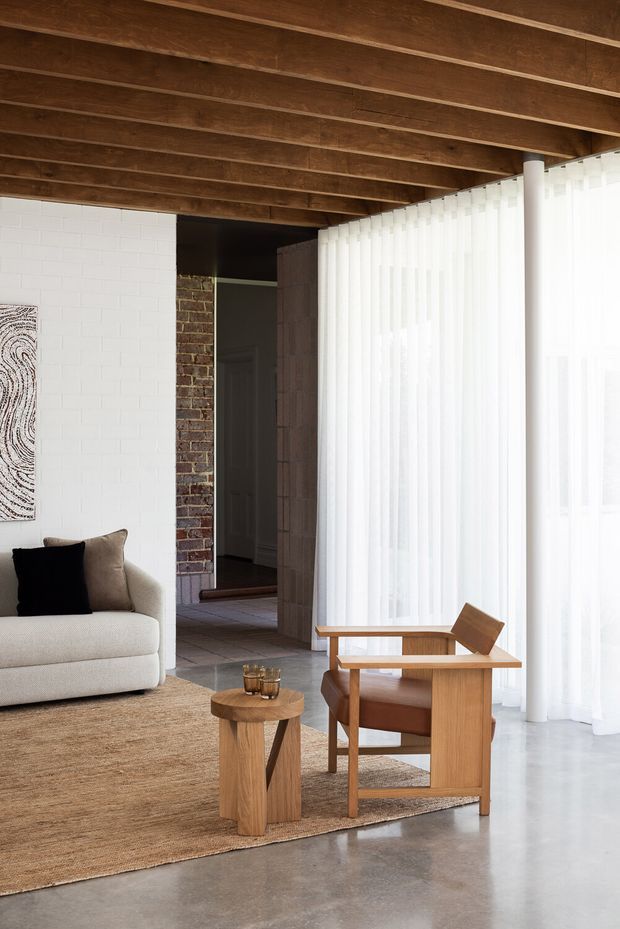 A refined living room with a warm exposed timber beam ceiling, white-painted brick walls, exposed brickwork, polished concrete flooring, and full-height sheer curtains. The room contains a light neutral sofa, a sculptural timber side table, a timber lounge chair, and a large natural-fibre rug. Abundant filtered daylight creates a calm, contemporary atmosphere.