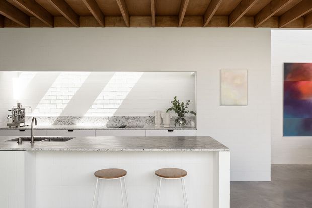 Bright minimalist kitchen with a large white island, pale stone benchtop, integrated sink and two timber-topped stools. A long horizontal interior window opens to a sunlit adjacent space, while exposed timber ceiling rafters add warmth and architectural character. White cabinetry and walls contrast with a polished grey concrete-like floor and colourful abstract artwork in the adjoining area.