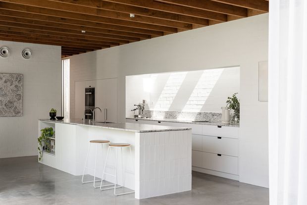 Bright open-plan kitchen with a large white island, integrated cabinetry, stone-look benchtops, a broad splashback, and two slim bar stools. The room features exposed warm timber ceiling beams, smooth white walls and joinery, polished grey concrete flooring, and abundant natural light.