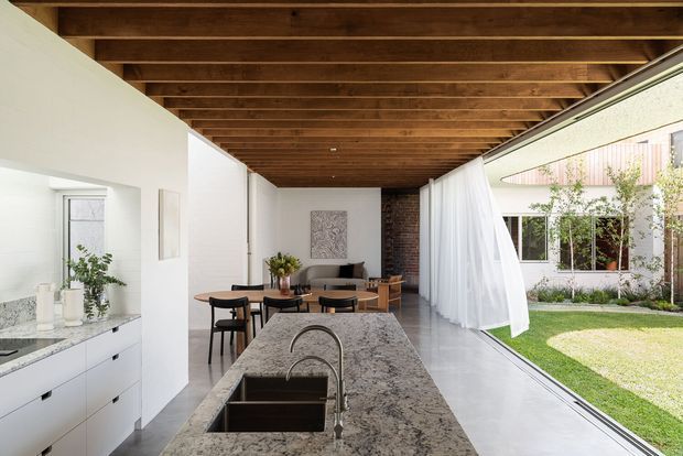 A bright open-plan kitchen and dining area arranged beneath a warm exposed timber-beam ceiling. The foreground features a long stone kitchen island with an integrated double sink and curved metal tap, while white cabinetry and a stone benchtop line the left wall. A dining table and chairs sit beyond the island, with a partially visible living area at the rear. Full-height glazed openings with sheer curtains connect the room to a landscaped courtyard, and a dark brick wall adds contrast at the back.