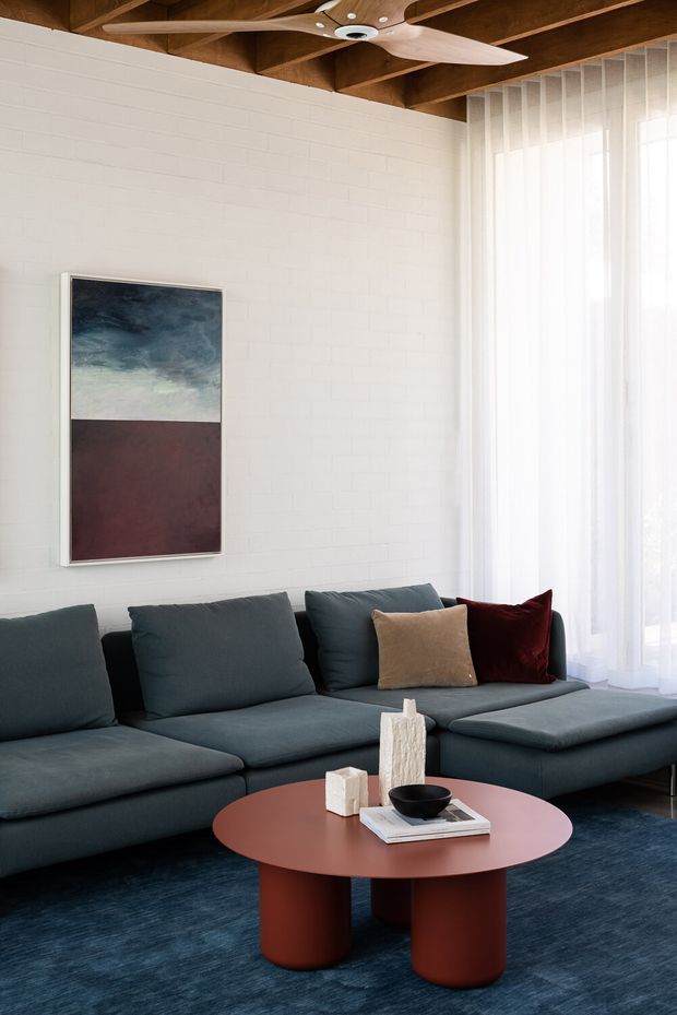 A styled living room with a large dark blue-grey sectional sofa, round rust-red coffee table, abstract artwork, and sheer floor-to-ceiling curtains. The room features a white painted brick wall, exposed timber ceiling beams, and a sculptural timber ceiling fan. A dark blue rug and restrained decorative objects create a contemporary, calm interior.