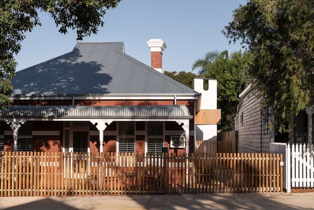 Street-facing view of a single-storey heritage-style house with a steep corrugated metal roof, exposed red-brick walls, decorative verandah detailing, a prominent brick chimney, and a timber picket fence. The facade combines traditional architectural elements with a contemporary white-and-timber chimney extension. Mature trees frame the property.