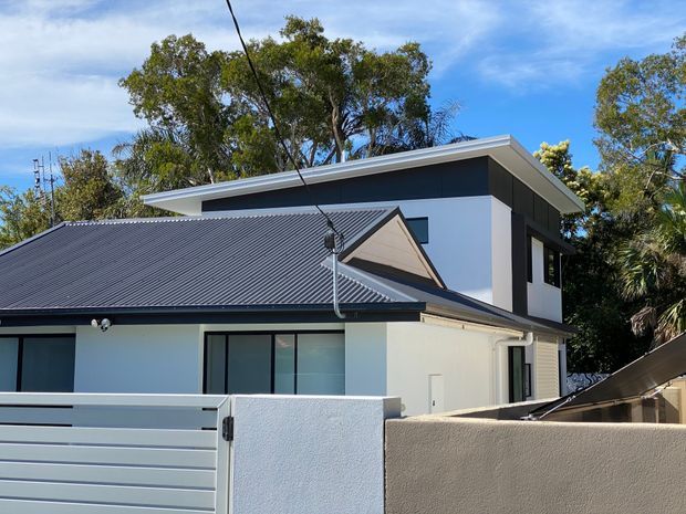 Exterior view of a contemporary two-storey residence with a low-pitched metal roof, contrasting white rendered walls and dark charcoal upper cladding. The home combines a broad main roof with a smaller gabled projection, dark-framed glazing, and a minimalist rendered boundary wall and gate. Mature trees surround the suburban property.