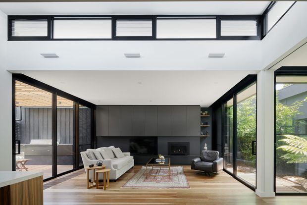 A bright contemporary living room with a double-height ceiling and high clerestory windows. The space features extensive black-framed floor-to-ceiling glazing, warm timber flooring, dark built-in cabinetry surrounding a fireplace and television, a white sectional sofa, a leather lounge chair, and a patterned area rug. Outdoor greenery and a courtyard are visible through the glazing.