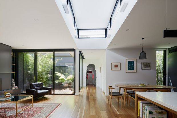 Bright open-plan residential interior combining a living room, dining area and kitchen. The space features warm timber flooring, extensive black-framed glazing, a large roof skylight, white walls and cabinetry, mid-century-inspired furniture, and a central hallway with an arched doorway.