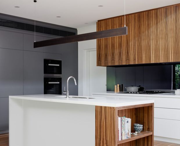 Contemporary kitchen with a large white island, integrated sink and tap, dark grey full-height cabinetry, built-in black appliances, and striking vertical-grain timber overhead cabinetry. A long dark linear pendant light spans the island, while an open timber display shelf adds warmth and storage.