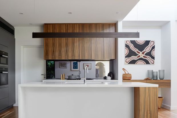 A contemporary open-plan kitchen featuring a large white island with integrated sink, dark built-in appliances, white cabinetry, and distinctive vertically grained timber overhead and lower joinery. A long dark horizontal pendant light spans the island, while a framed abstract artwork, timber bench, decorative ceramics, and woven basket add warmth. The room has a bright, clean, refined appearance with recessed ceiling lighting.