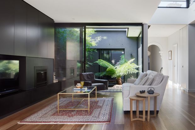 A bright contemporary living room with a dark built-in media wall and fireplace, large black-framed glazing opening to a planted courtyard, a pale sofa and armchair, glass-and-brass coffee table, patterned rug, and timber flooring. A skylight and high ceiling add natural light, while an arched opening leads toward the adjacent circulation area.