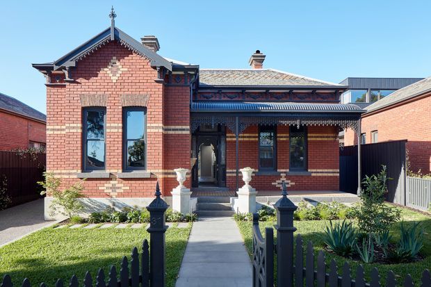 Front exterior view of a restored single-storey red-brick heritage house, featuring ornate Victorian detailing, a steep gabled front section, decorative bargeboards and verandah trim, tall narrow windows, a metal roof, front steps, and a landscaped garden enclosed by a decorative picket fence. A contemporary addition is visible behind the original facade.