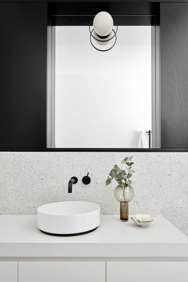 A refined contemporary bathroom vanity photographed straight on. The composition features a white vessel basin on a pale stone benchtop, matte black wall-mounted tapware, a speckled light terrazzo or engineered-stone splashback, dark timber-framed mirrored cabinetry above, and a minimalist spherical wall light. A small vase and dish provide subtle styling.