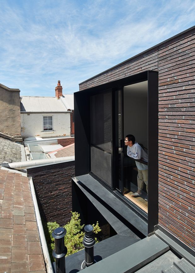 Exterior architectural photograph looking across a dense urban rooftop context toward a contemporary brick-clad building extension. The dark, recessed façade features a tall glazed opening and projecting black metal sill or awning, with a person visible inside. Adjacent older brick and rendered buildings, corrugated roofing, rooftop services, and a small planted lightwell are visible. Bright daylight and blue sky emphasize the contrast between the historic surroundings and the modern addition.