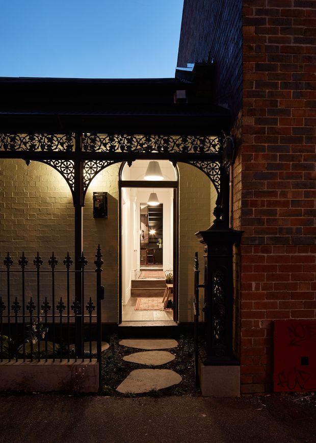 A dusk exterior view of a narrow residential entry courtyard. A decorative black wrought-iron gate and ornate lacework veranda frame a lit doorway leading into a long interior hallway. The surrounding facade combines exposed red brick with pale painted brick walls, a dark roof, traditional ironwork, and stepping-stone paving.