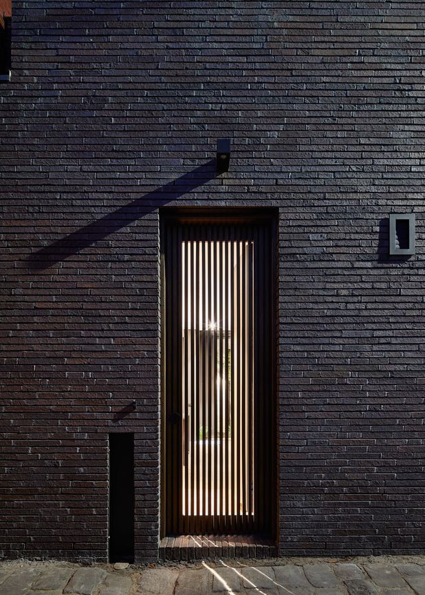 Close-up exterior view of a contemporary dark brick facade with a tall narrow entrance door screened by vertical timber slats. Strong sunlight casts diagonal shadows across the textured masonry, with a small wall-mounted light and paved threshold visible.