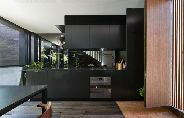A contemporary dark-toned kitchen with extensive matte black cabinetry, integrated stainless-steel oven, dark benchtops, and a reflective splashback or mirrored rear wall. Floor-to-ceiling glazing and vertical timber screening bring natural light into the space, while indoor plants and a timber dining table add warmth. A staircase and adjacent living area are visible through the reflective surfaces and opening.