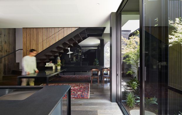 A refined open-plan contemporary interior viewed from a dark stone kitchen island, with a dining and living area extending beneath a dramatic sculptural staircase. Full-height black-framed glazing opens to a planted courtyard, while vertical timber slats, dark cabinetry, timber flooring, and a richly patterned rug add contrast and warmth.