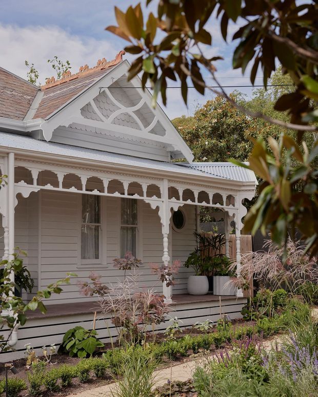 Exterior view of a finely detailed historic timber cottage, featuring pale weatherboard cladding, an ornate gabled roof, decorative fretwork veranda, turned posts, scalloped trim, and a landscaped front garden. The image has a soft, warm heritage character with the house framed by foreground foliage.