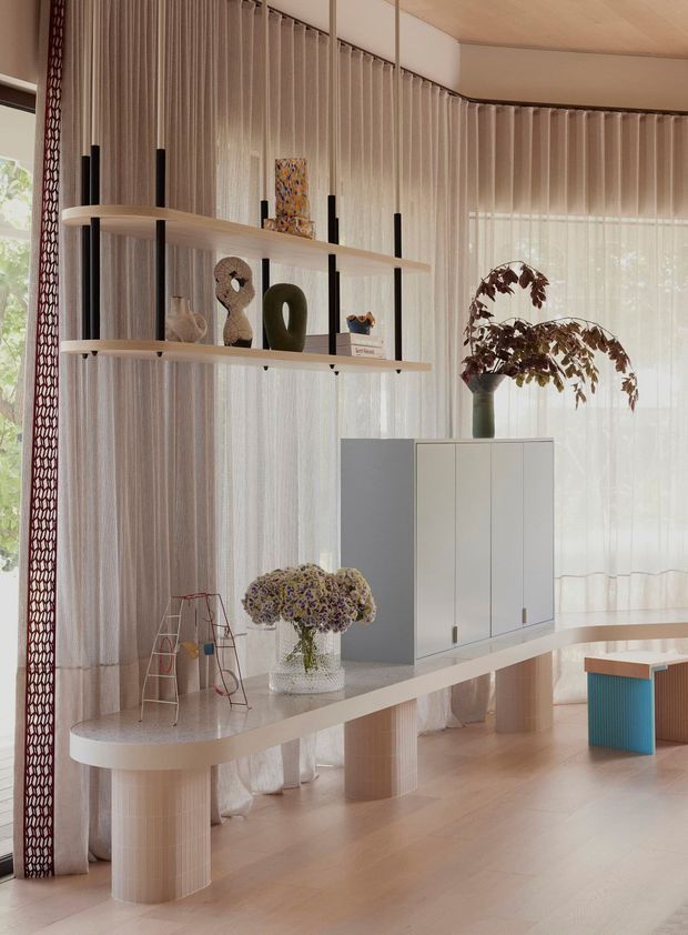 A refined contemporary interior featuring a sculptural curved console or display table with cylindrical supports, floating open shelves, and a white built-in cabinet. Sheer floor-to-ceiling curtains soften the room, while decorative objects, artwork, and dried floral arrangements add warmth. The space uses pale timber flooring, light neutral surfaces, and subtle patterned screens or curtains, creating an airy, curated living area.