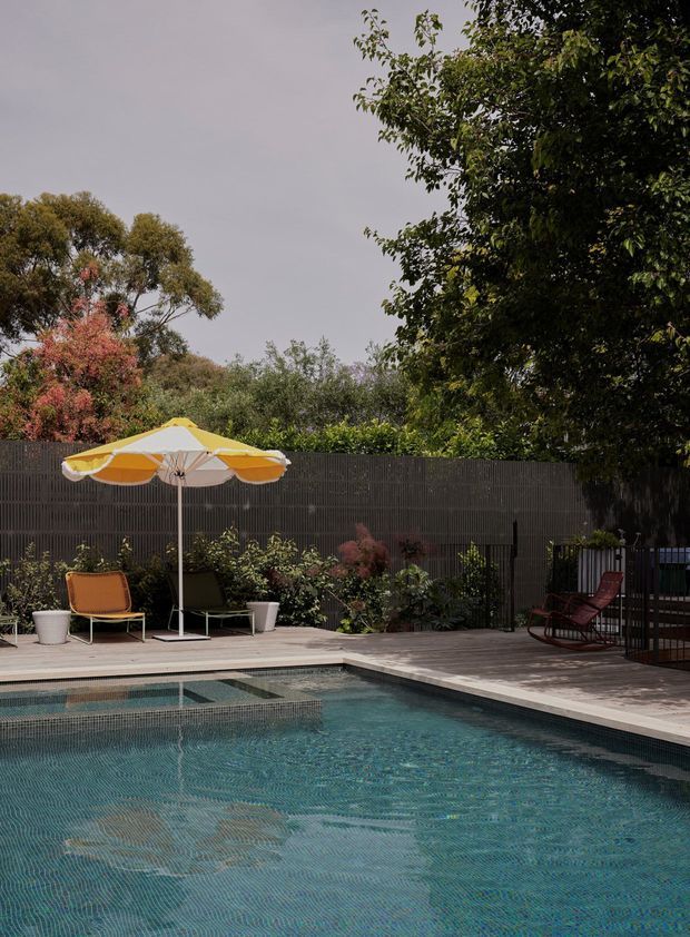 A landscaped residential pool area with a turquoise tiled swimming pool in the foreground, surrounded by a timber deck, dark vertical privacy fencing, potted plants, outdoor lounge chairs, and a yellow-and-white patio umbrella. Mature trees and dense planting create a private garden setting.
