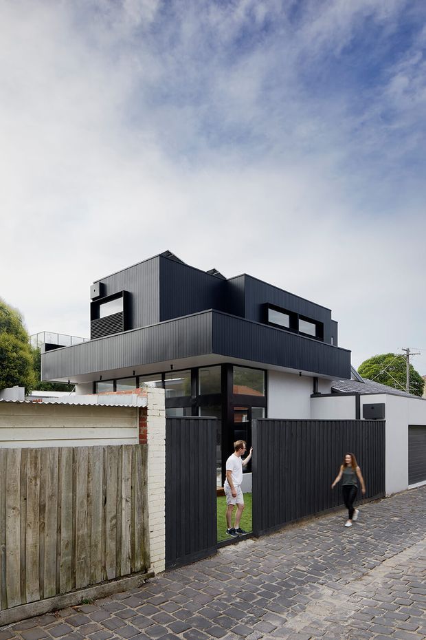 Street-facing view of a contemporary multi-storey residence with a bold dark vertically ribbed cladding exterior, projecting upper levels, large dark-framed windows, a high boundary fence and gate, and a cobbled external walkway. Older masonry and timber fencing sit alongside the newer structure, with mature vegetation and neighbouring buildings visible around it.