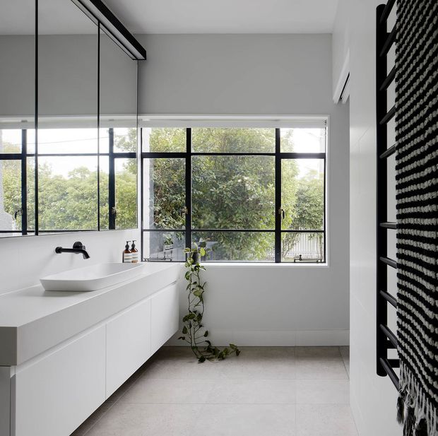Bright contemporary bathroom with a long floating white vanity, integrated basin and wall-mounted black tapware. Large black-framed windows provide abundant natural light and views of greenery, while a black heated towel rail with textured towels occupies the right wall. Pale tiled flooring, white walls, and a trailing indoor plant create a clean, airy interior.
