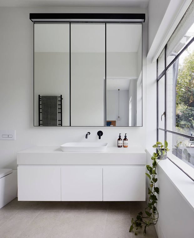 Bright, contemporary bathroom with a wall-mounted white vanity, integrated storage, white vessel basin, matte black tapware, and a large three-panel mirrored cabinet. Full-height black-framed windows bring in abundant natural light, with a trailing indoor plant beside the vanity. Large-format grey floor tiles and minimal white finishes create a clean, refined appearance.