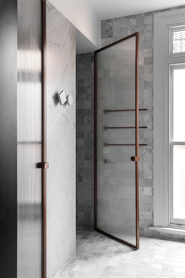 A refined contemporary bathroom featuring grey veined stone-look wall and floor tiles, a walk-in shower enclosed by ribbed translucent glass panels in dark timber or bronze-toned frames, and matching horizontal shower niches or shelving. A tall white-framed window provides natural light, while a pale stone-clad partition and dark fixtures create a restrained, high-contrast palette.